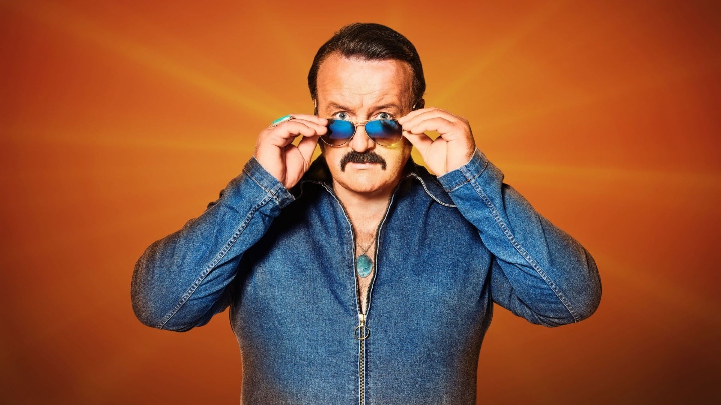 A man wearing 80s clothing with slick hair and aviator sunglasses stood against an orange background