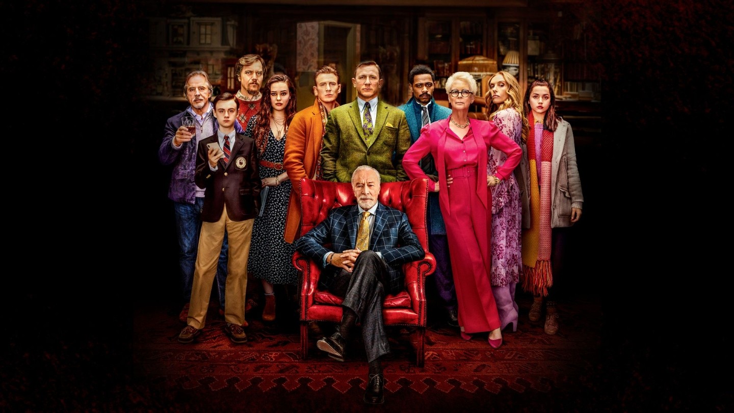 Group of nine people in colorful formal and semi-formal clothing standing behind a person seated in a red armchair, set in an elegant room with dark wood furniture and patterned carpet.