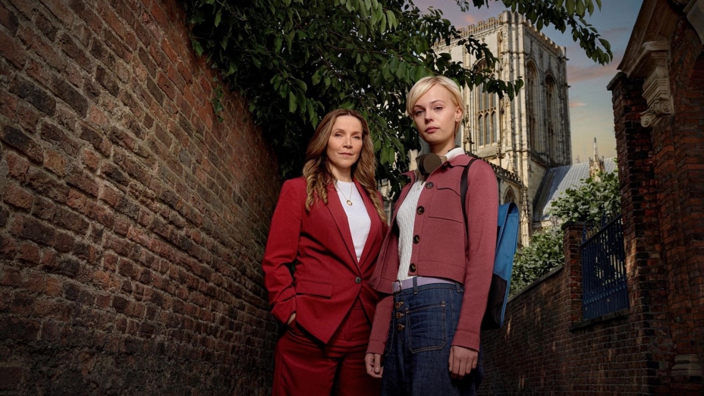 Promotional image for the second season of the British crime drama Patience, showing two people standing in a narrow brick alley with greenery overhead and a large historic cathedral in the background. One person wears a red suit, and the other wears a pink cardigan with a blue backpack. 