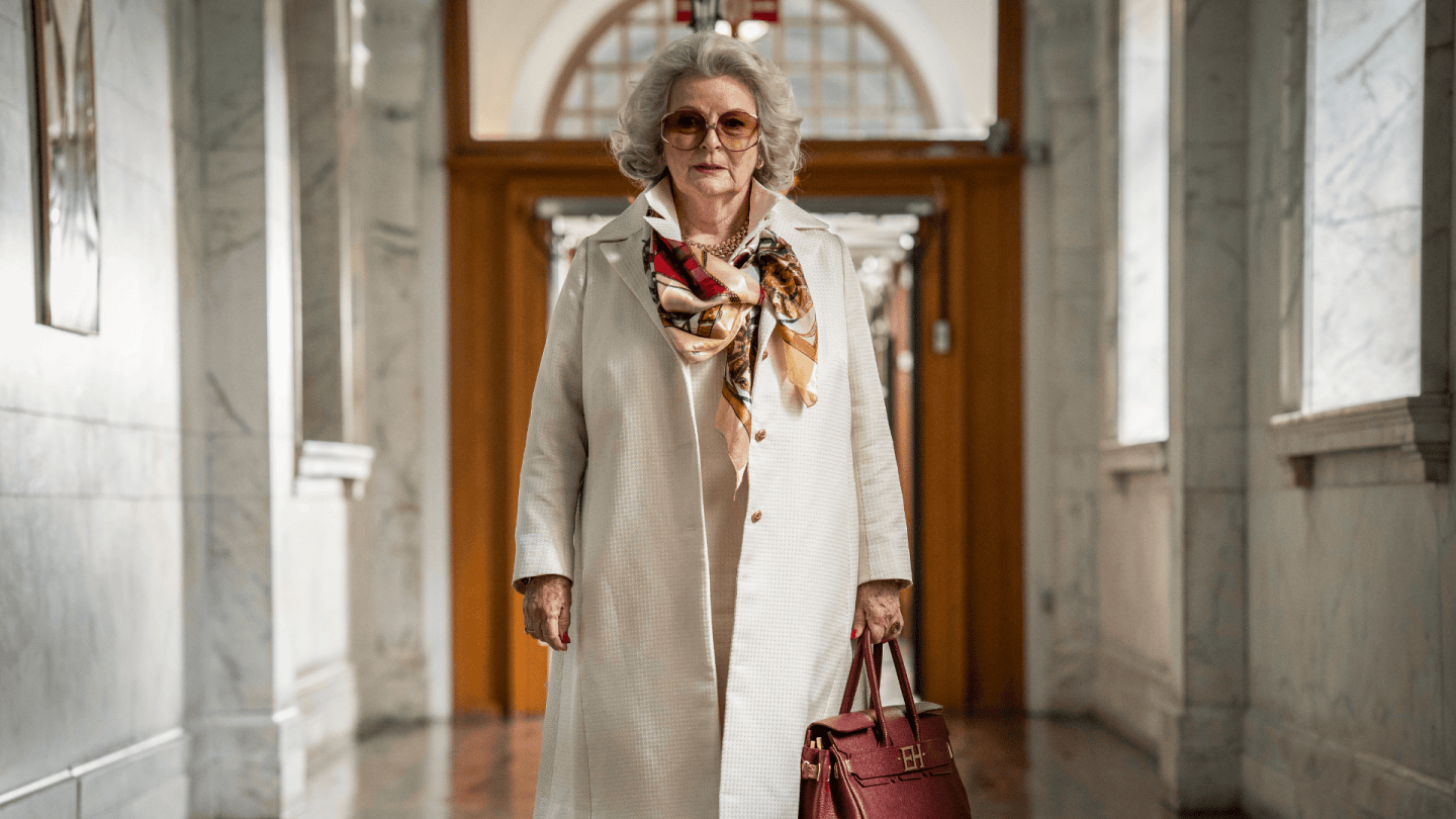 Person walking through a grand marble hallway wearing a light coat, patterned scarf and carrying a red handbag.