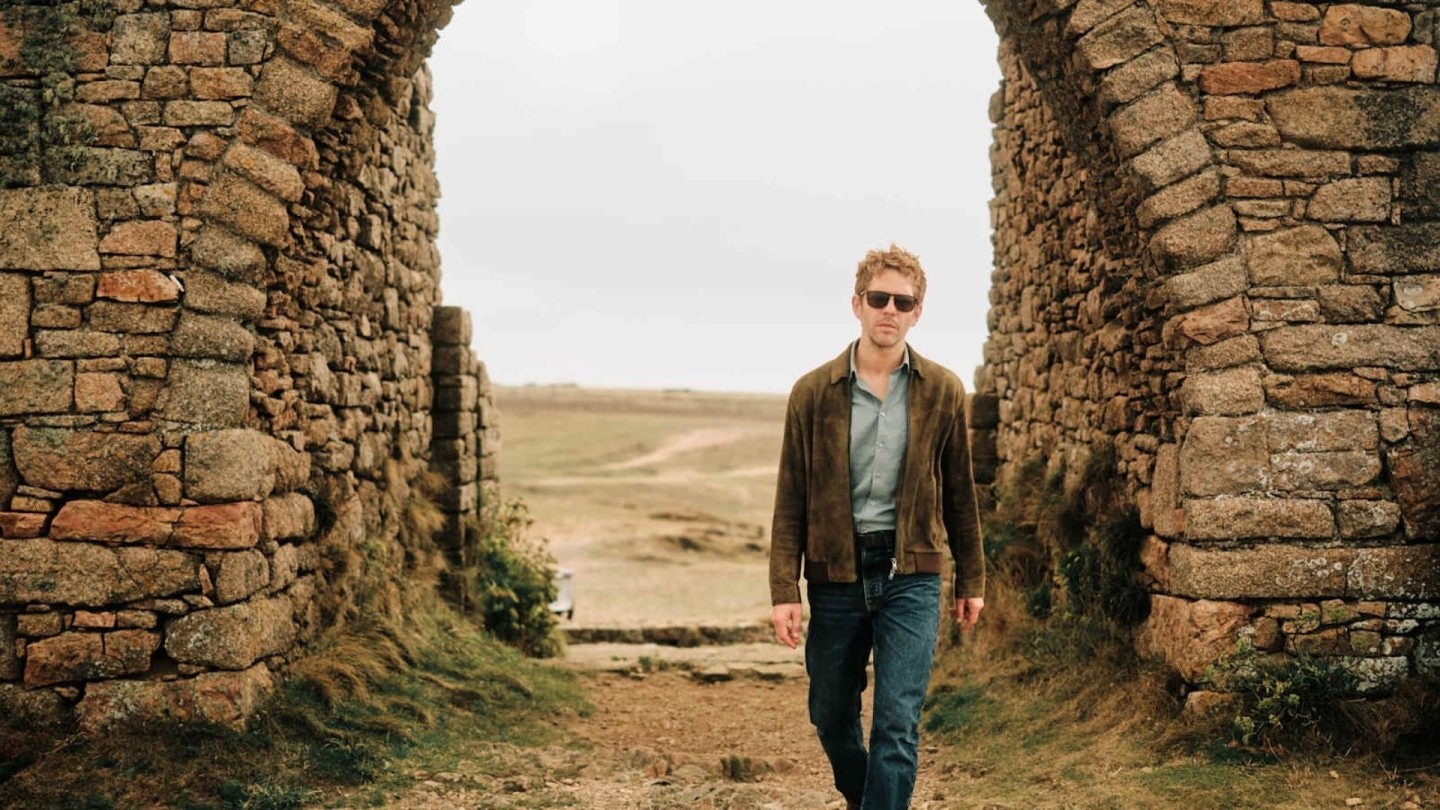Damian Moloney as Jim Bergerac wearing a tanned jacket and blue jeans walking towards the camera with a countryside backdrop 