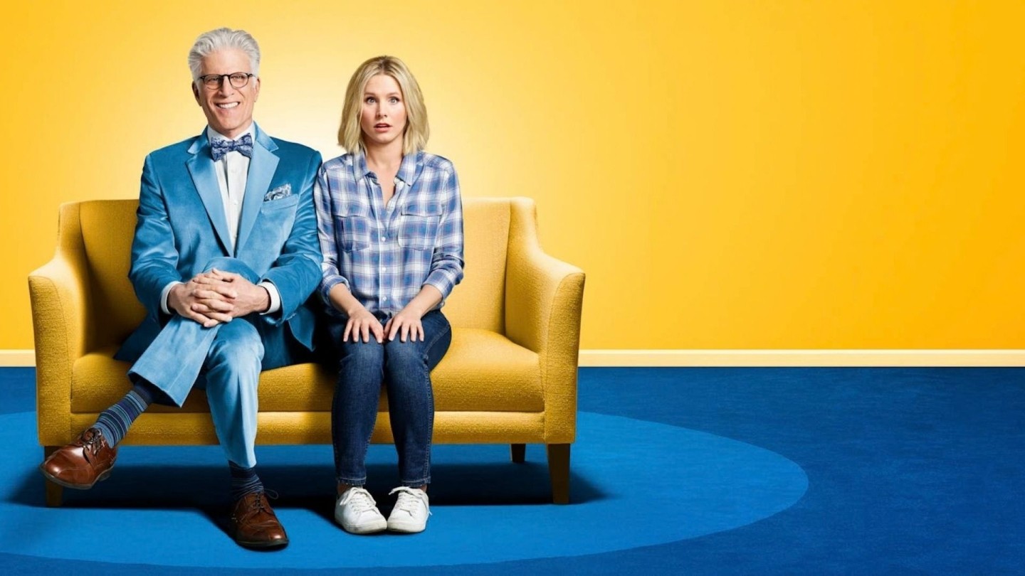 A man and a women wearing blue sat on a yellow sofa on blue carpet against a yellow background. Key art image for The Good Place.
