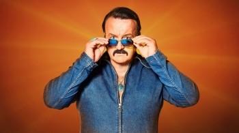 A man wearing 80s clothing with slick hair and aviator sunglasses stood against an orange background