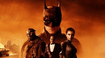 Poster image for the 2022 film The Batman, featuring Batman in the background with a fiery orange sky and rain. In the foreground, three characters stand together, including one in a leather outfit and another wearing glasses and a heavy coat. A burning car and city buildings are visible in the distance. 