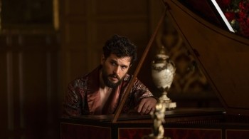 A still image from season 4 of the US drama series Industry, showing a person in an ornate patterned robe leaning on a grand piano in a richly decorated room with dark wood panelling and a decorative vase in the foreground.