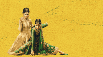 Two individuals wearing ornate traditional South Asian attire against a vibrant yellow textured background, one dressed in an elegant cream and gold embroidered outfit standing gracefully, while the other in a rich green and gold embellished ensemble poses dramatically on the floor.