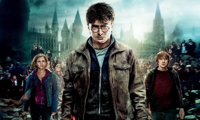 Daniel Radcliffe stood in an a shabby jacket in front of the backdrop of Hogwarts castle with Rupert Grint and Emma Watson stood behind him
