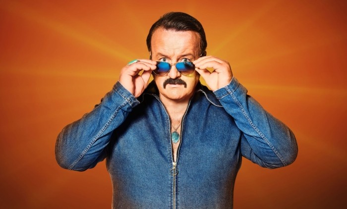 A man wearing 80s clothing with slick hair and aviator sunglasses stood against an orange background