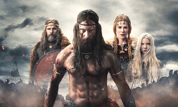 A viking with brown hair shirtless and with tattoos stood in front of the image with two female vikings and a male stood behind him against a stormy background in the movie The Northman