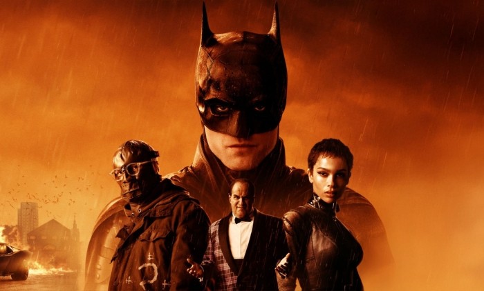 Poster image for the 2022 film The Batman, featuring Batman in the background with a fiery orange sky and rain. In the foreground, three characters stand together, including one in a leather outfit and another wearing glasses and a heavy coat. A burning car and city buildings are visible in the distance. 