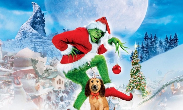 Green character dressed as Santa holding a red ornament in a snowy Whoville setting with a decorated Christmas tree and full moon.