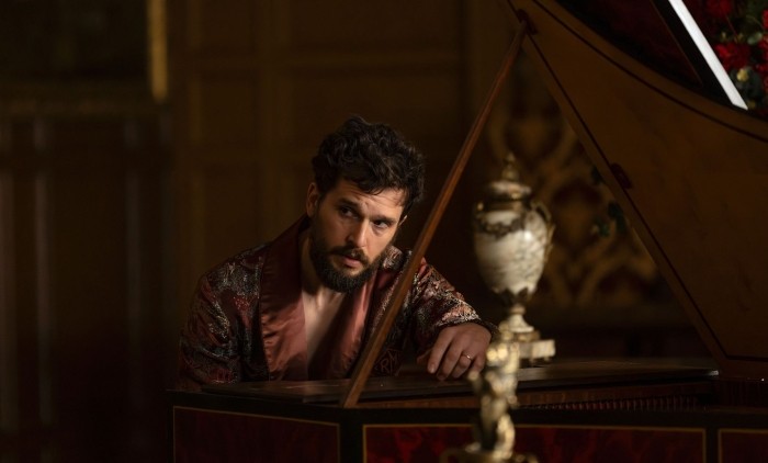 A still image from season 4 of the US drama series Industry, showing a person in an ornate patterned robe leaning on a grand piano in a richly decorated room with dark wood panelling and a decorative vase in the foreground.