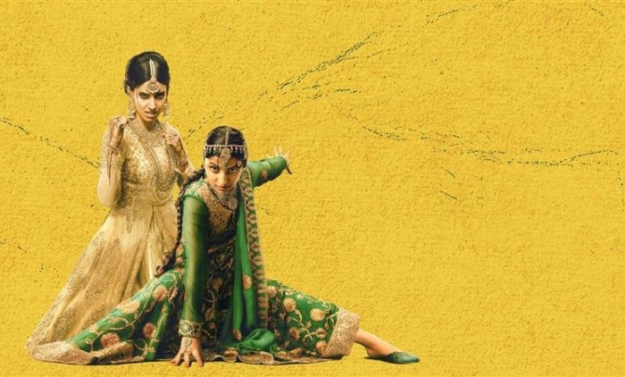 Two individuals wearing ornate traditional South Asian attire against a vibrant yellow textured background, one dressed in an elegant cream and gold embroidered outfit standing gracefully, while the other in a rich green and gold embellished ensemble poses dramatically on the floor.