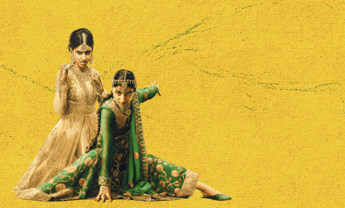Two individuals wearing ornate traditional South Asian attire against a vibrant yellow textured background, one dressed in an elegant cream and gold embroidered outfit standing gracefully, while the other in a rich green and gold embellished ensemble poses dramatically on the floor.