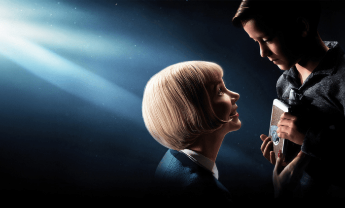 Two people standing closely under dramatic studio lighting, with one holding a vintage camera against a dark, atmospheric background.