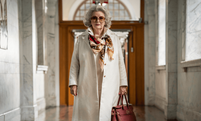 Person walking through a grand marble hallway wearing a light coat, patterned scarf and carrying a red handbag.