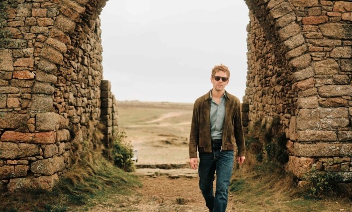 Damian Moloney as Jim Bergerac wearing a tanned jacket and blue jeans walking towards the camera with a countryside backdrop 