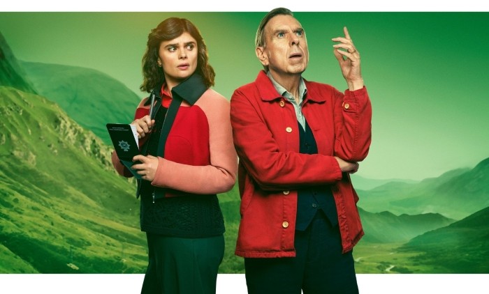 Promotional image for the drama series Death Valley, showing two people standing back to back in a mountainous landscape, one holding a notebook, both wearing red‑toned jackets against a green valley backdrop.