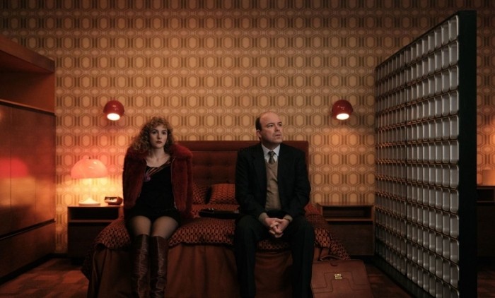 Promotional image for Honey showing two characters seated on a bed in a retro-style hotel room with warm orange lighting, patterned wallpaper and vintage furnishings, reflecting the drama’s period setting and intimate character-driven story.