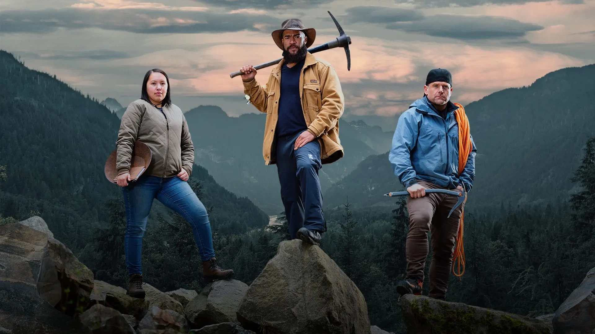 A man stood on a rocj above a valley holding a pickaxe behind him with a women stood just below on the left and a slightly older man stood on the right