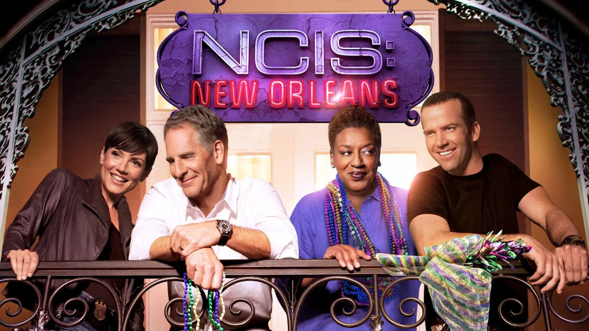 The cast of NCIS New Orleans stood on a balcony wearing early noughties clothing smiling