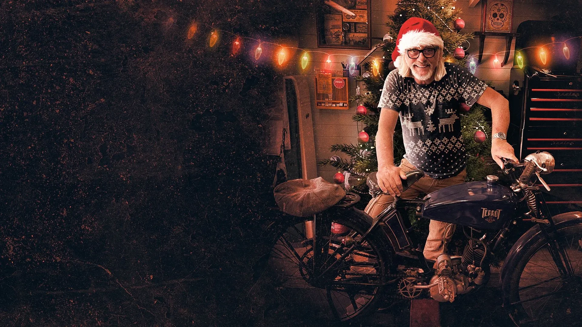 A man wearing a Christmas hat holding on old vintage bike with fairy lights behind him