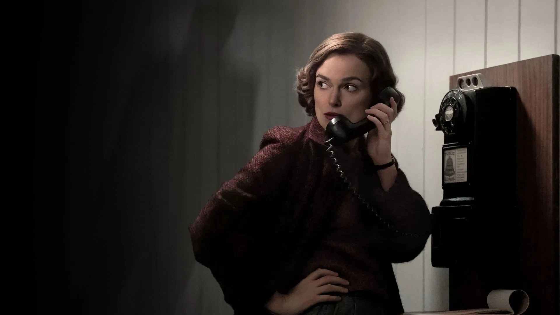 Person standing by a vintage rotary payphone, holding the receiver, against a dimly lit wall.