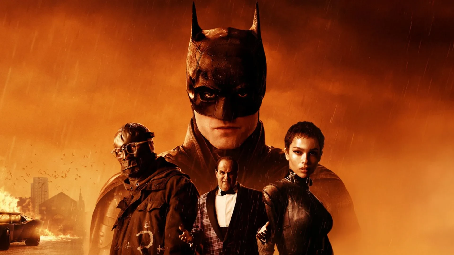 Poster image for the 2022 film The Batman, featuring Batman in the background with a fiery orange sky and rain. In the foreground, three characters stand together, including one in a leather outfit and another wearing glasses and a heavy coat. A burning car and city buildings are visible in the distance. 