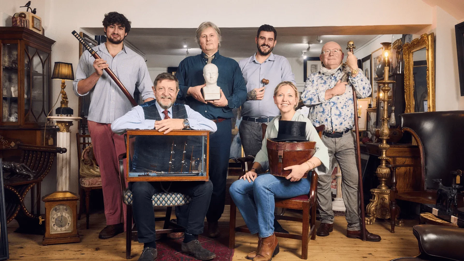 Group of antique experts posing with vintage items in a traditional antiques shop filled with classic furniture and collectibles.