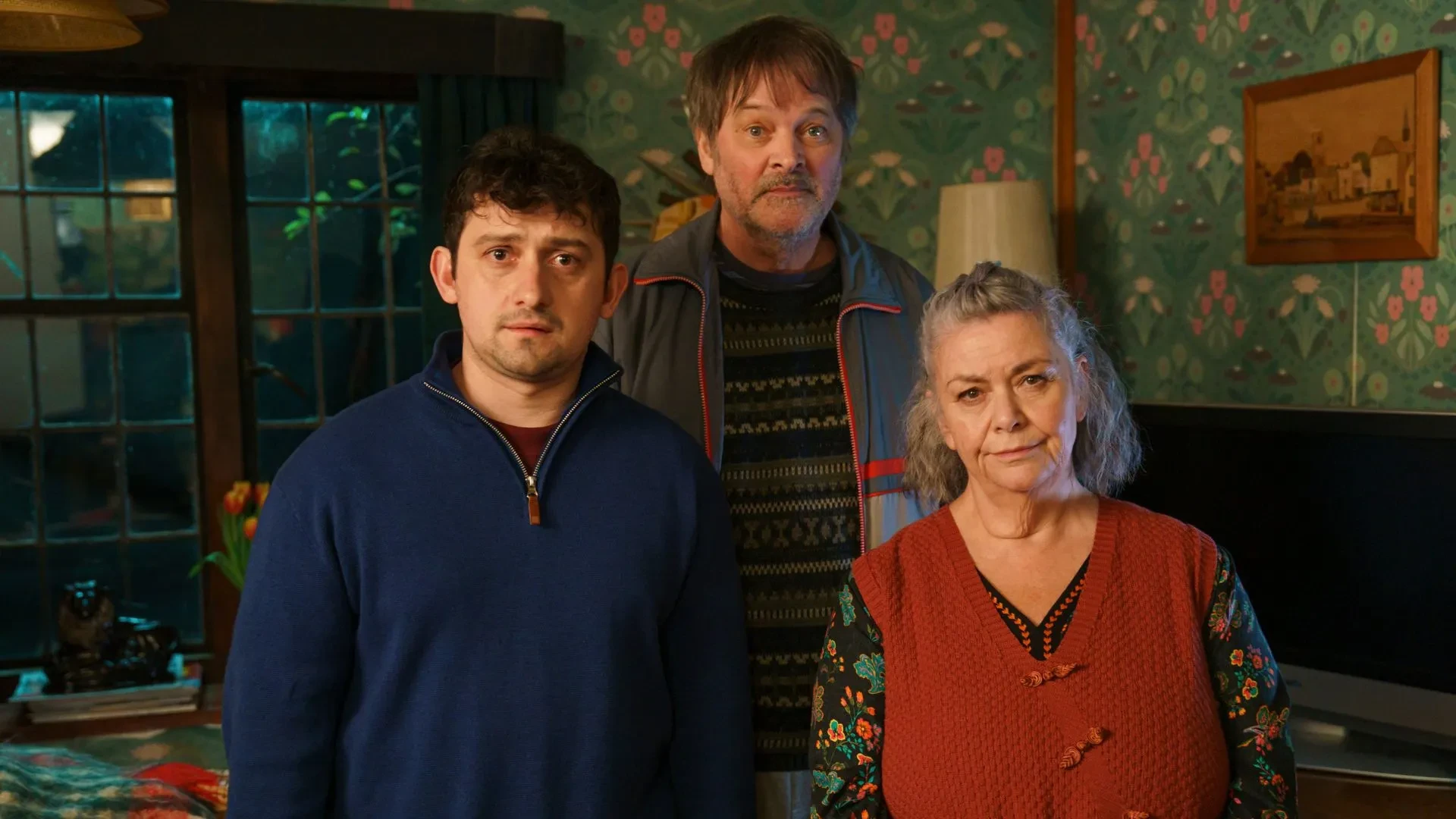 A still image from the British comedy drama series Can You Keep a Secret?, showing three people standing in a cosy living room with patterned green wallpaper, a large window, a television, and a lamp. One person wears a blue zip-up top, another a dark patterned jumper with a jacket, and the third a red knitted vest over a floral shirt.