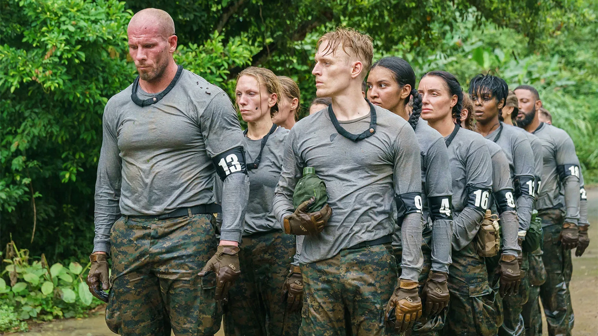 Promotional image from the eighth series of the British reality competition Celebrity SAS: Who Dares Wins, showing a line of contestants in grey long-sleeved tops and camouflage trousers standing outdoors in a jungle setting. They wear black gloves and numbered armbands, and one person holds a green water bottle.