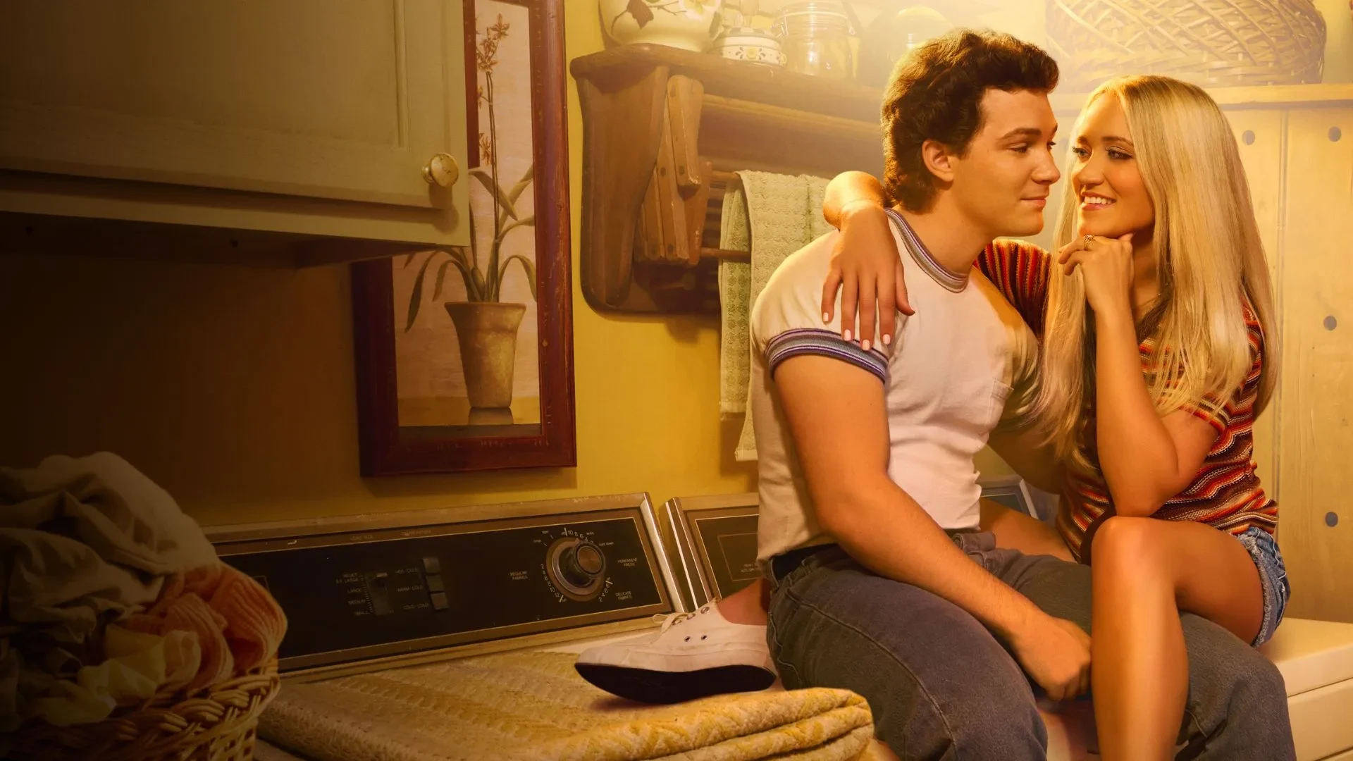 Promotional image showing two people sitting closely together on top of a washing machine in a cosy laundry room. The person on the left is wearing a white T-shirt with striped sleeves and jeans, while the person on the right is dressed in a colourful striped top and denim shorts. The background features wooden cabinets, a wicker basket, folded towels, and a framed floral artwork, creating a warm, homely atmosphere.