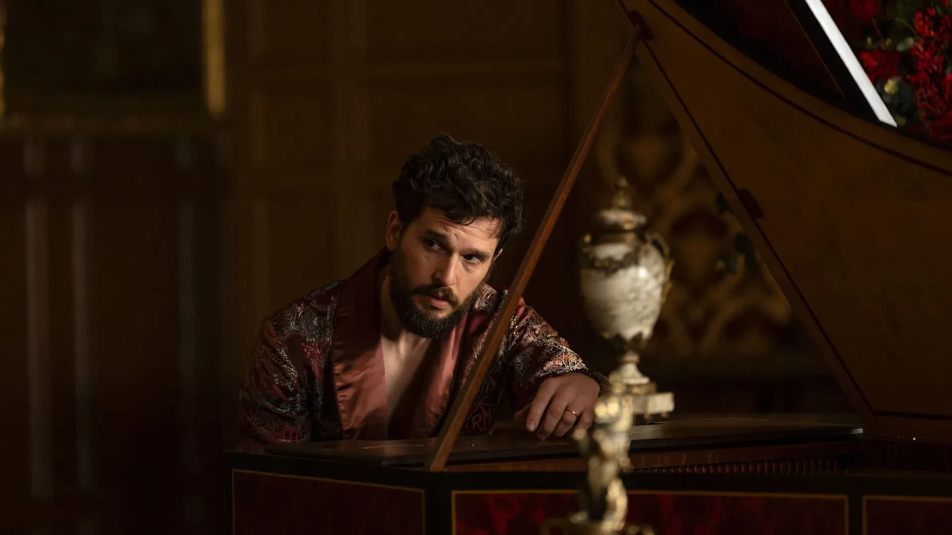 A still image from season 4 of the US drama series Industry, showing a person in an ornate patterned robe leaning on a grand piano in a richly decorated room with dark wood panelling and a decorative vase in the foreground.
