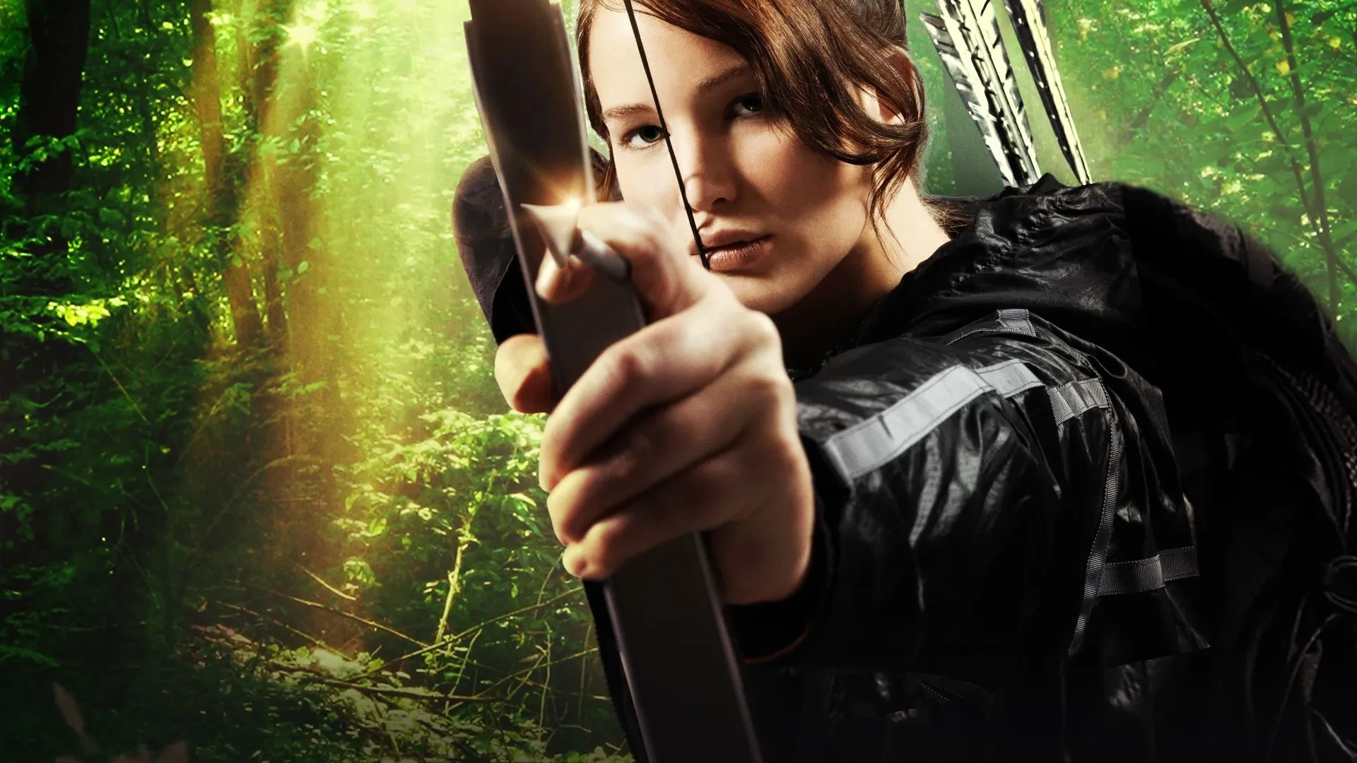 Jennifer Lawrence in a dark jacket draws a bow and arrow in a sunlit forest, with green foliage and light beams in the background.