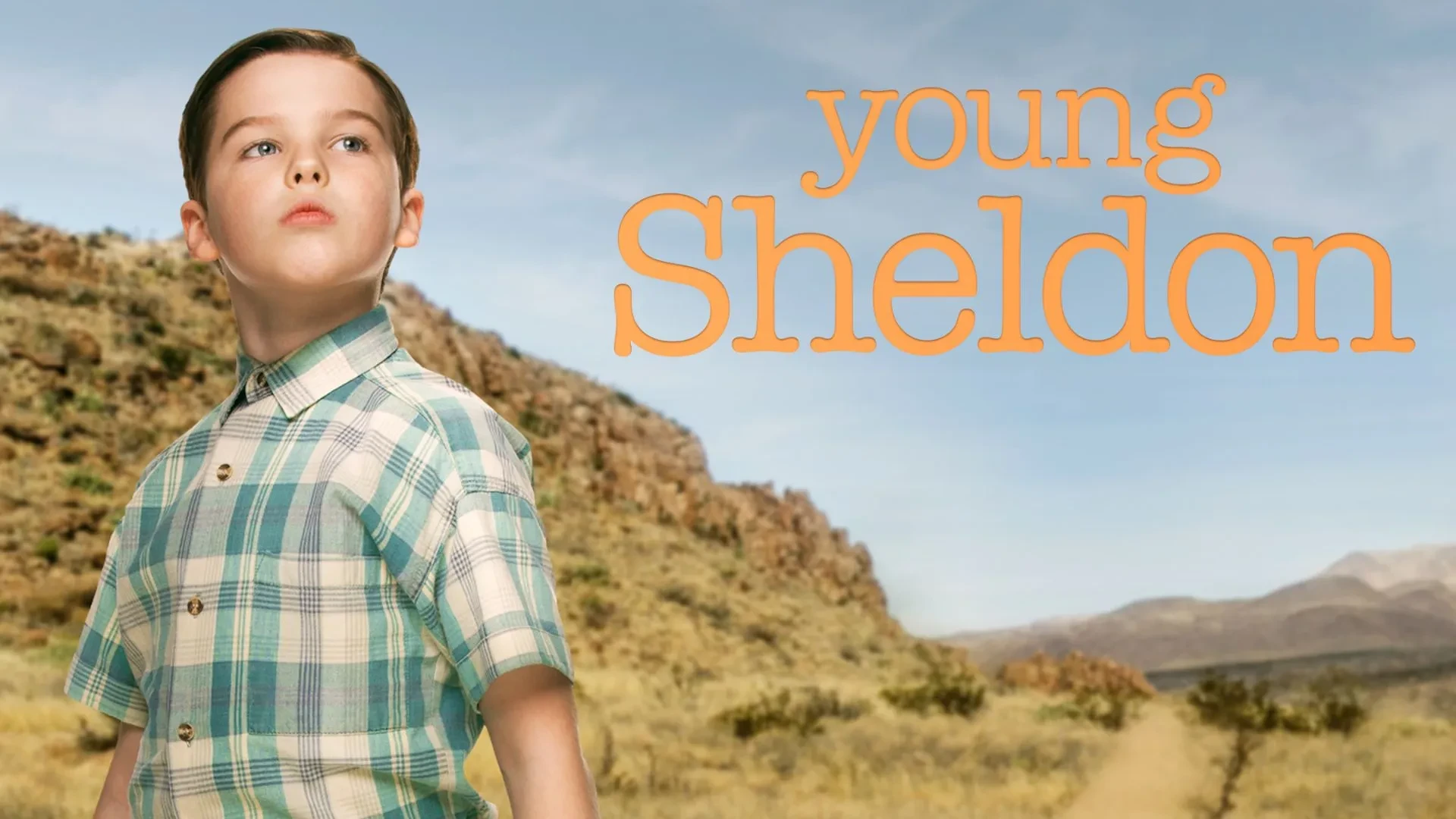 Promotional image for Young Sheldon featuring a child wearing a green and white checked short-sleeve shirt, standing outdoors in a desert landscape with rocky hills and a clear blue sky. The series title ‘Young Sheldon’ appears in large orange text on the right side of the image.