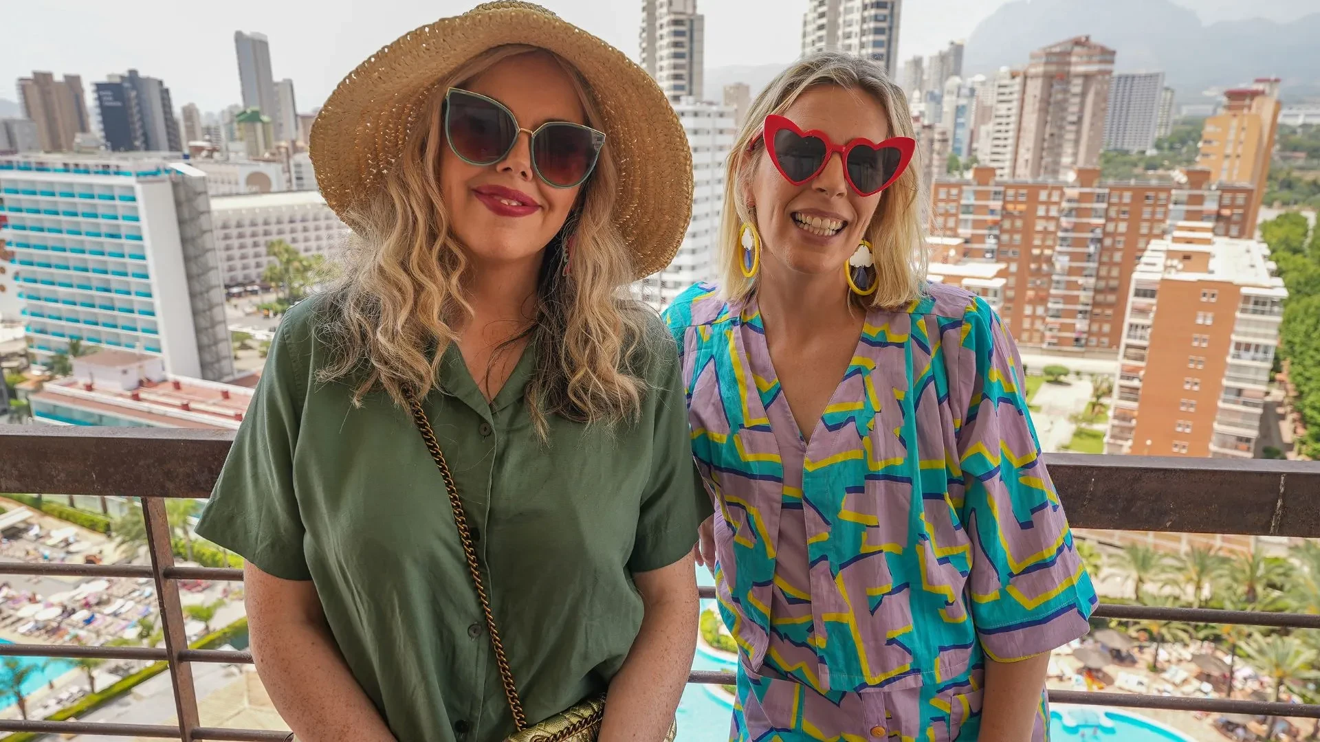 Two people standing on a balcony overlooking a cityscape with high-rise buildings, hotels, and a swimming pool below. One person is wearing a wide-brimmed straw hat and a loose green shirt with a gold chain strap bag, while the other is dressed in a colourful patterned top with bold geometric shapes in turquoise, purple, and yellow, accessorised with large round earrings and red sunglasses. The background features palm trees and mountains in the distance under a bright, clear sky.