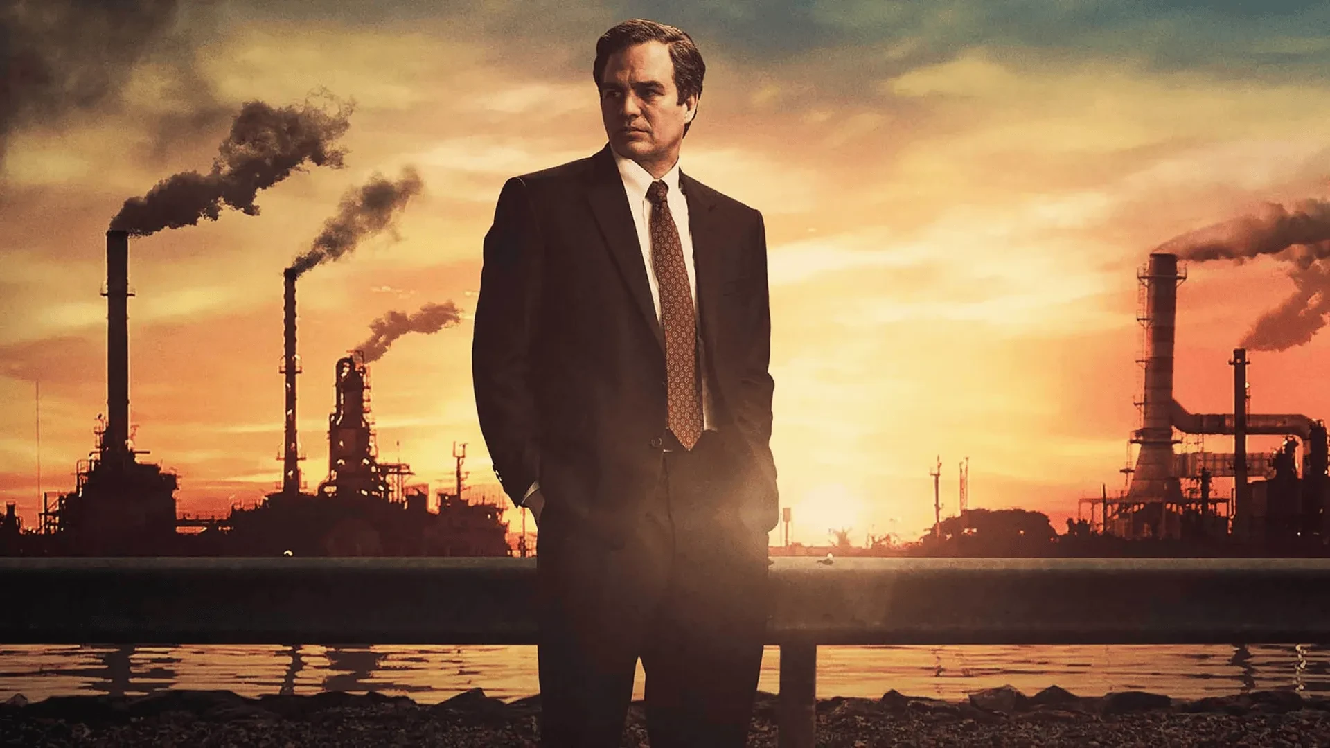 Person in a business suit standing in front of a large industrial plant at sunset, with smokestacks rising into an orange sky.
