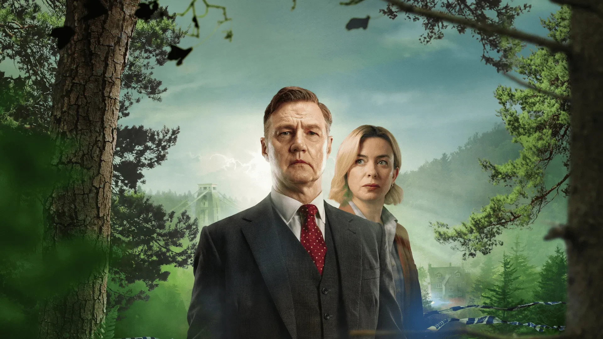 Two individuals standing in a forest clearing with scenic hills, trees and a distant suspension bridge behind them.