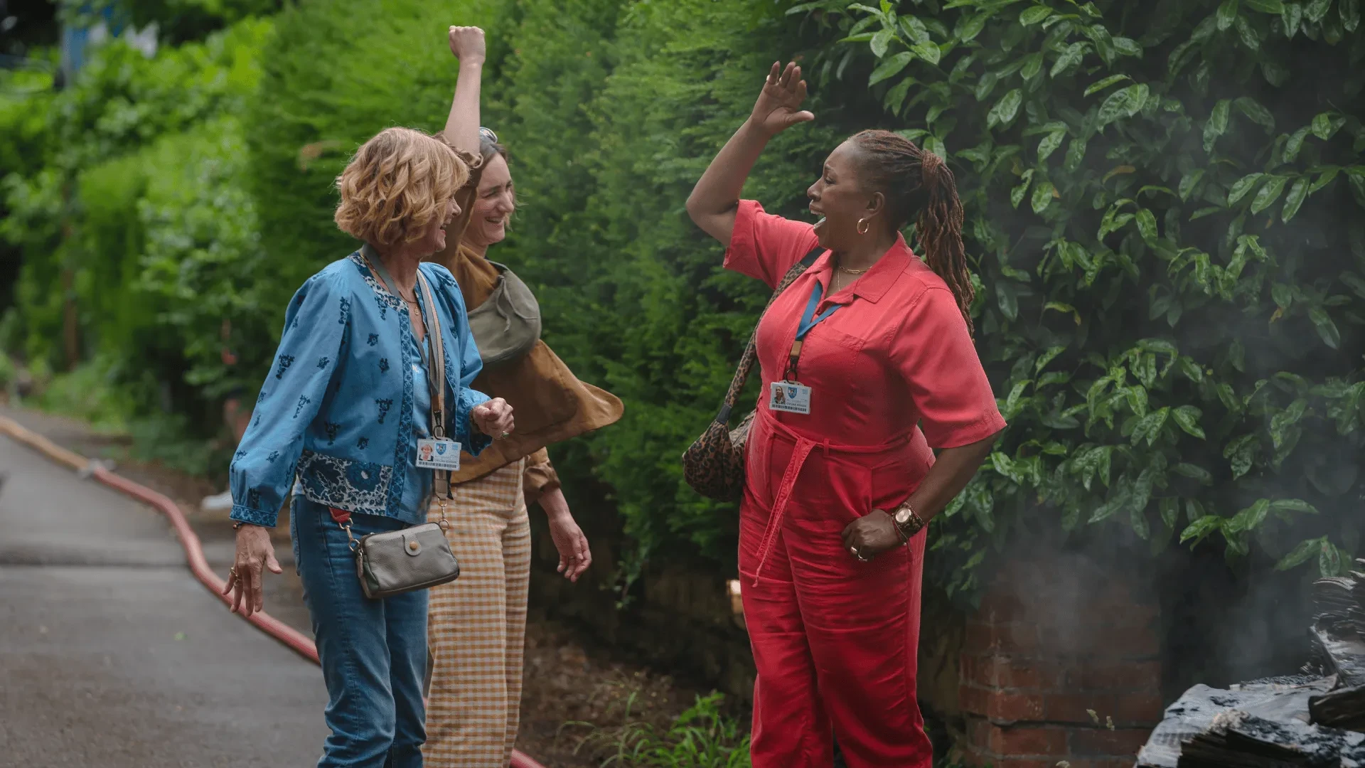 Three people standing on a path beside tall green hedges, gesturing toward something out of view during an outdoor discussion