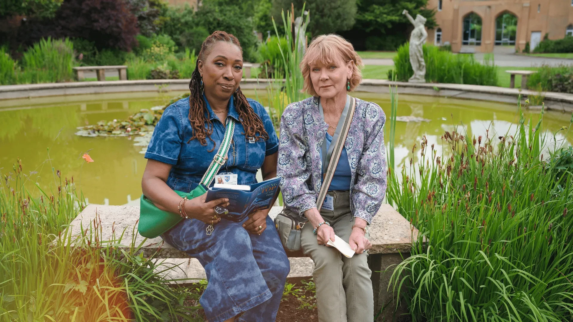 Two people sitting on a stone bench beside a pond filled with water lilies and surrounded by lush greenery