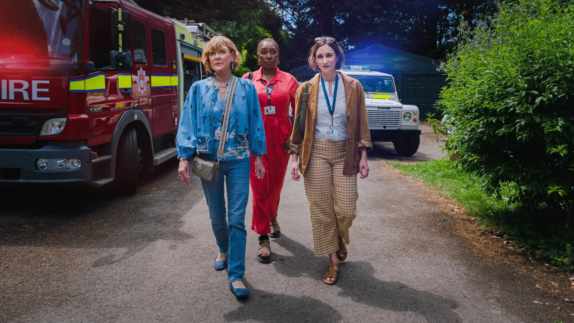 Three people walking down a path near emergency vehicles including a fire engine and an off‑road rescue vehicle.