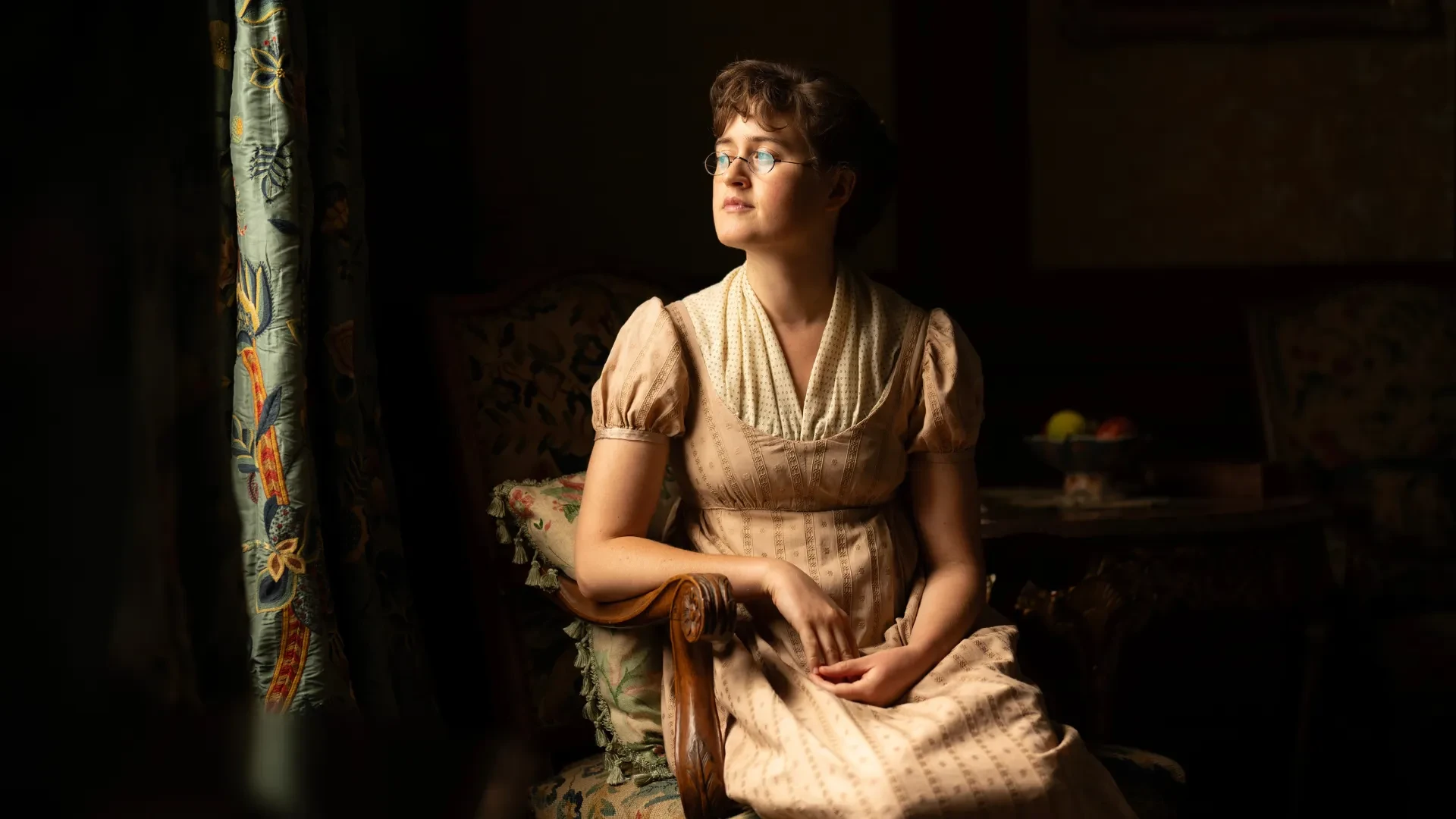 Person seated in an antique chair wearing a period costume in a dimly lit, traditionally decorated room