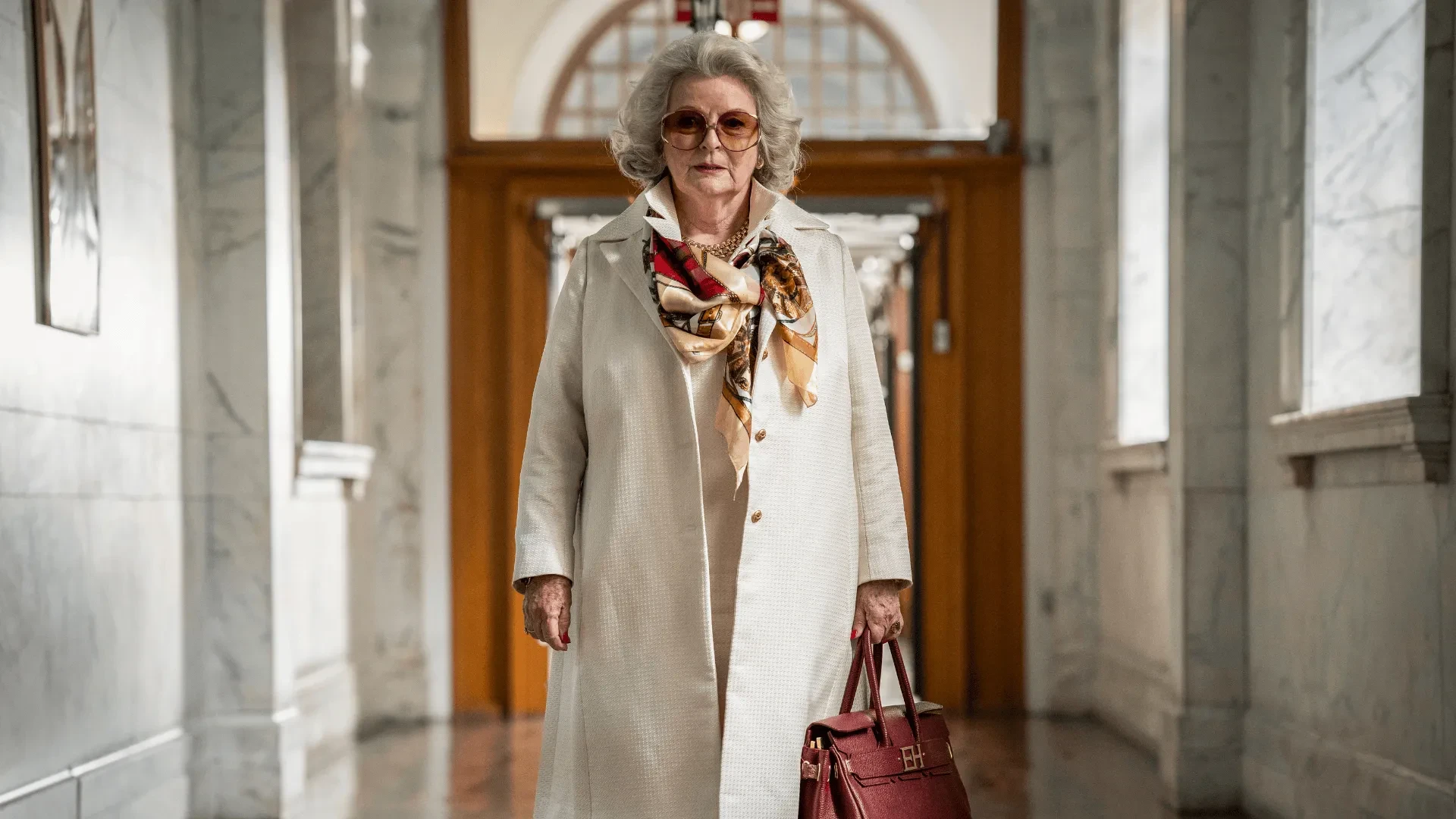 Person walking through a grand marble hallway wearing a light coat, patterned scarf and carrying a red handbag.