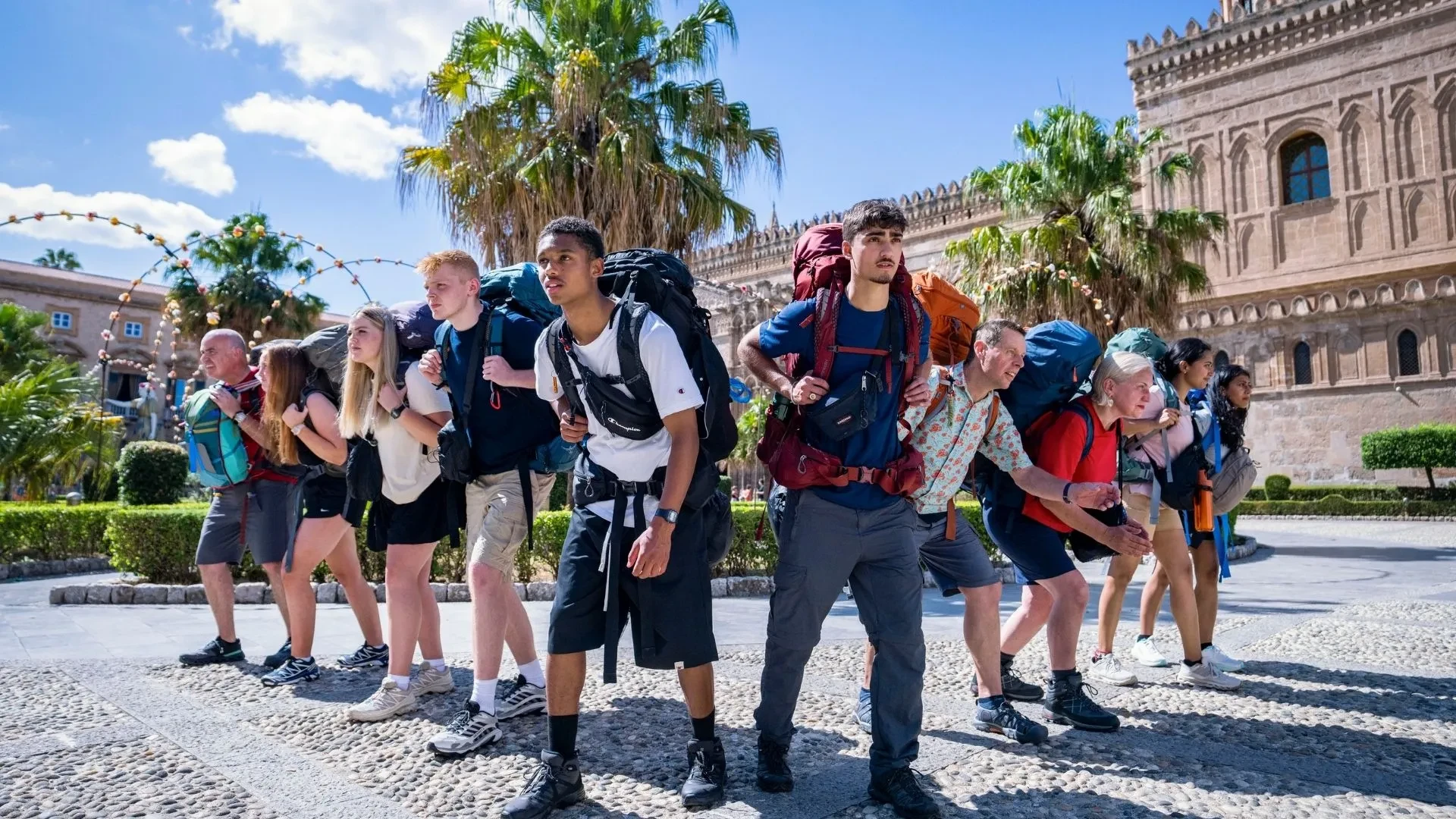 Contestants in Race Across the World wearing summer hiking clothing with huge backpacks stood in front of an Italian looking building, posed to run