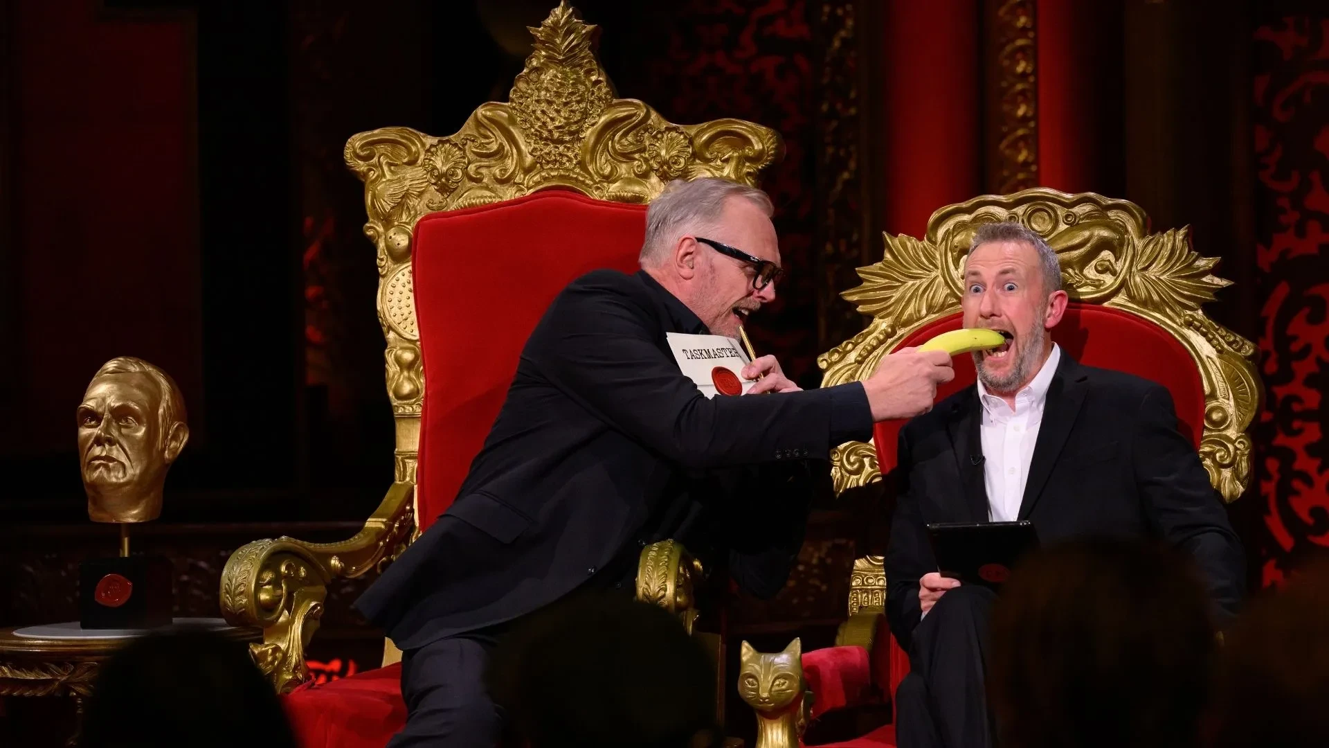 Greg Davies and Little Alex Horne sat on Taskmaster chairs with Greg holding a banana