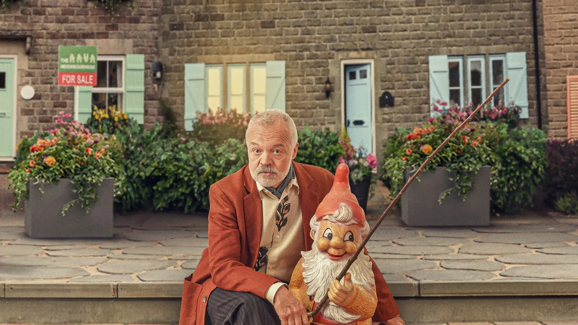Graham Norton with a red jacket sat on a bench outside a nice house holding a gnome for the new reality series The Neighbourhood