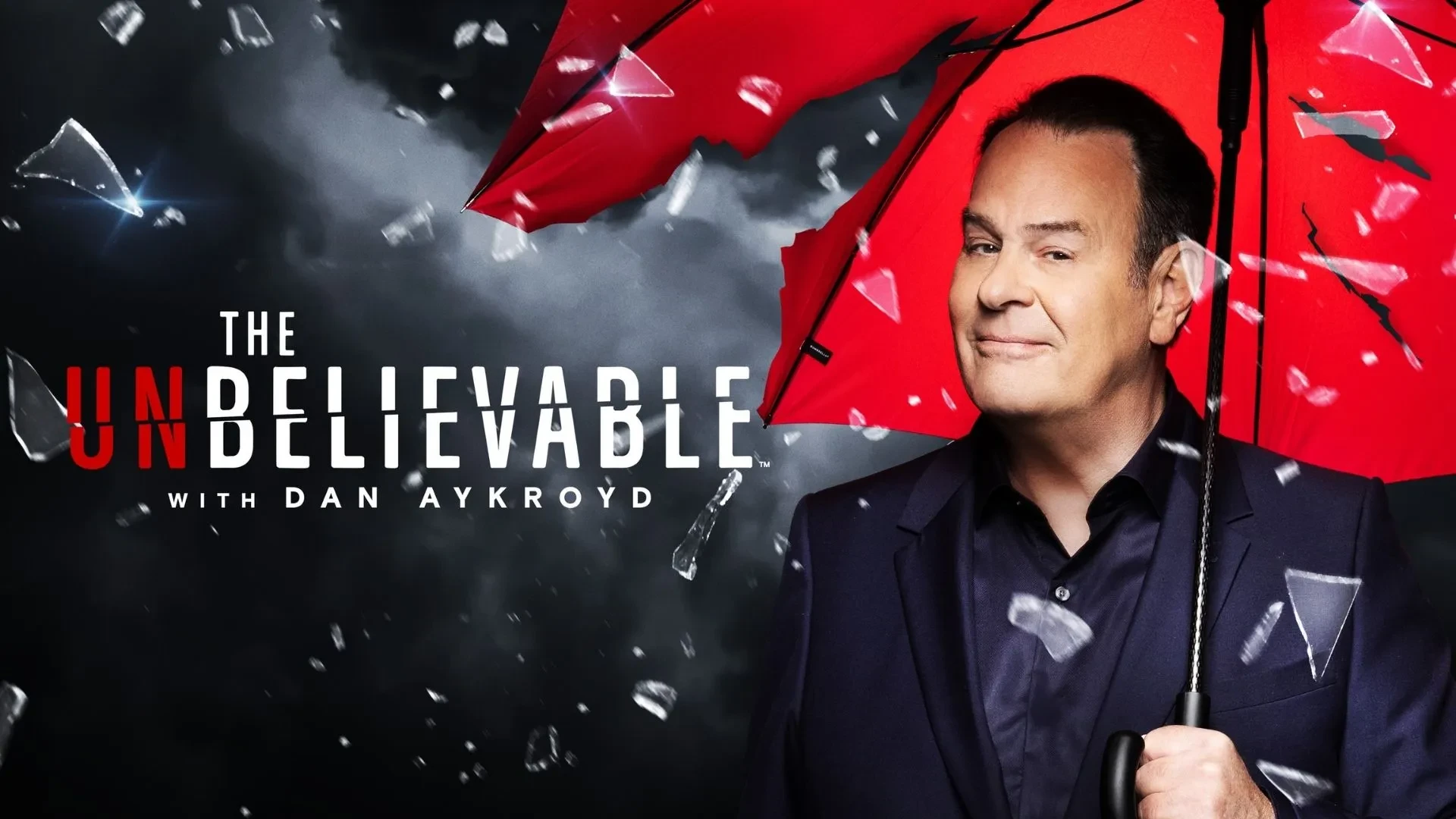 Dan Aykroyd stood under a red umbrella against a black backdrop that reads  "The Unbelievable with Dan Aykroyd"