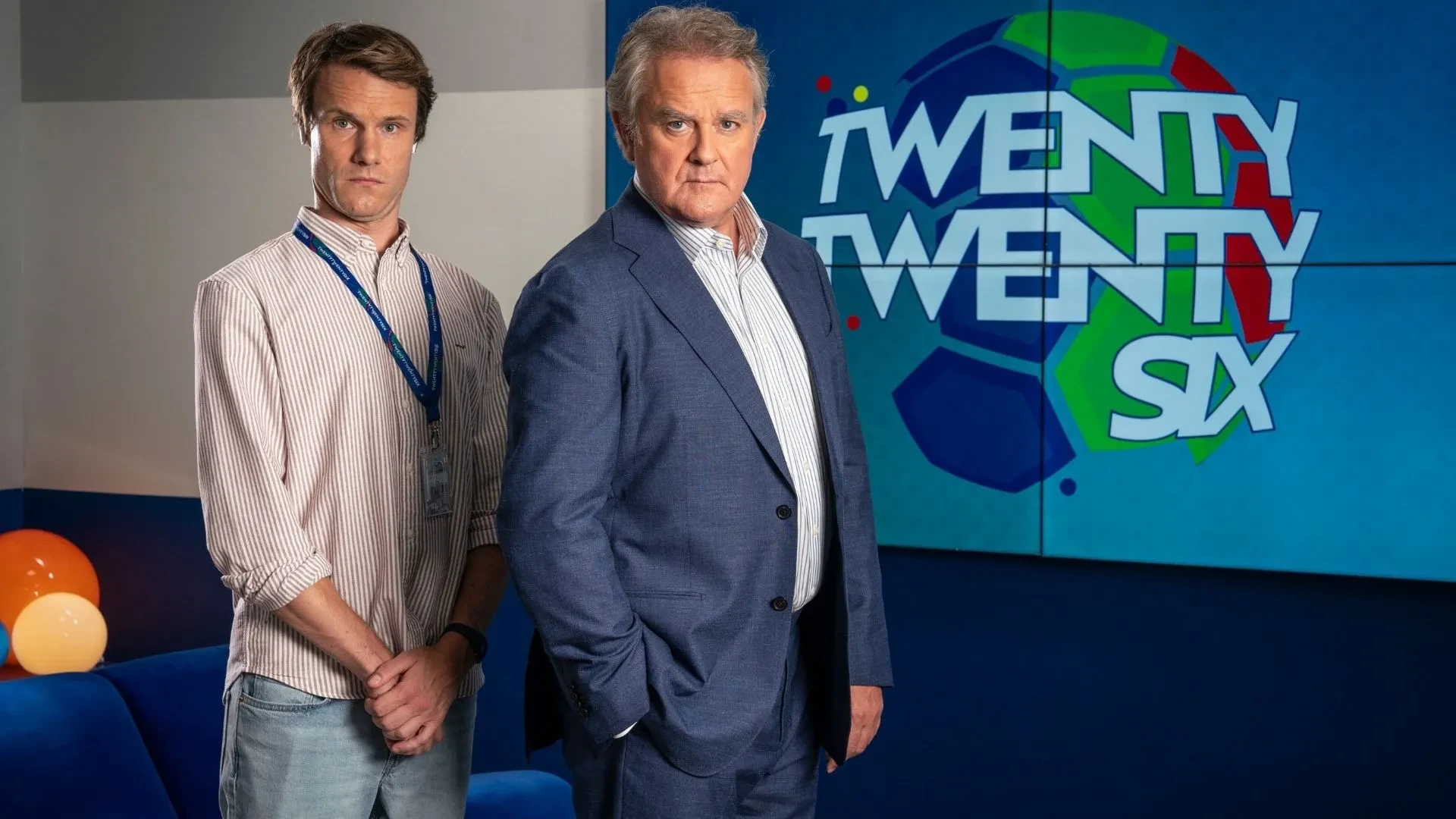 Hugh Bonneville and Hugh Skinner in BBCs comedy Twenty Twenty Six stood in smart clothing against a sign labelled "twenty twenty six"
