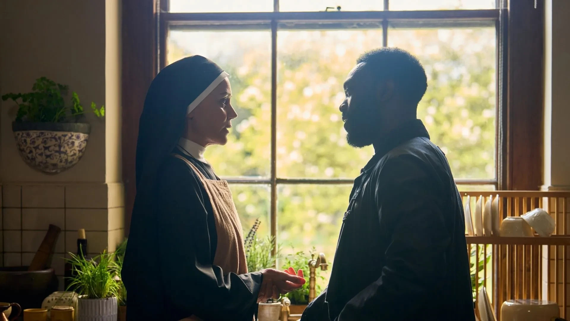 Promotional image for the drama Falling, showing two people standing face to face in a softly lit kitchen interior, with a large window, houseplants, dishes, and cooking utensils visible in the background.