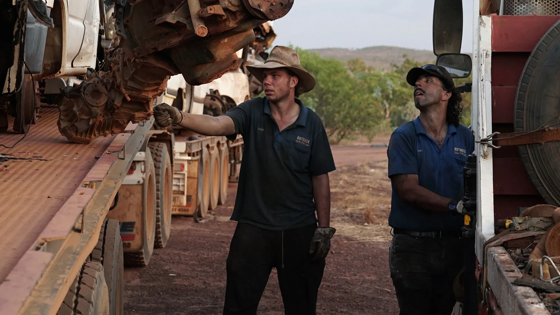 Two mechanics stood between lorries holding old rusty cars, examining them
